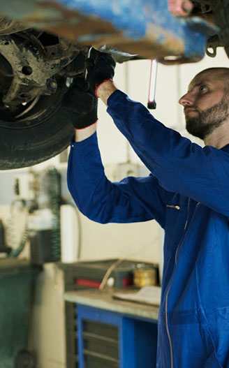A mechanic in a blue jumpsuit works underneath a vehicle, using a tool to adjust or repair the car's components in a workshop.