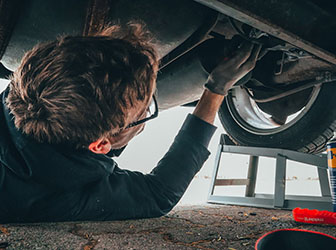A person in glasses lies under a vehicle, using tools to inspect or repair the exhaust system.