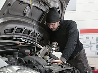 A person works on a car engine under the hood, using a flashlight to inspect components in a workshop setting.